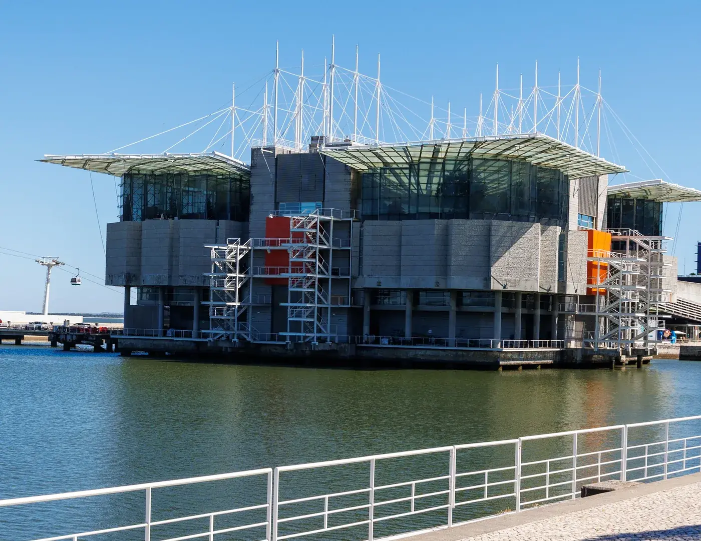 The Oceanário de Lisboa's grey, glass-topped pavilion standing on its pier in the lagoon at Parque das Nações, a cable-car cabin passing behind it